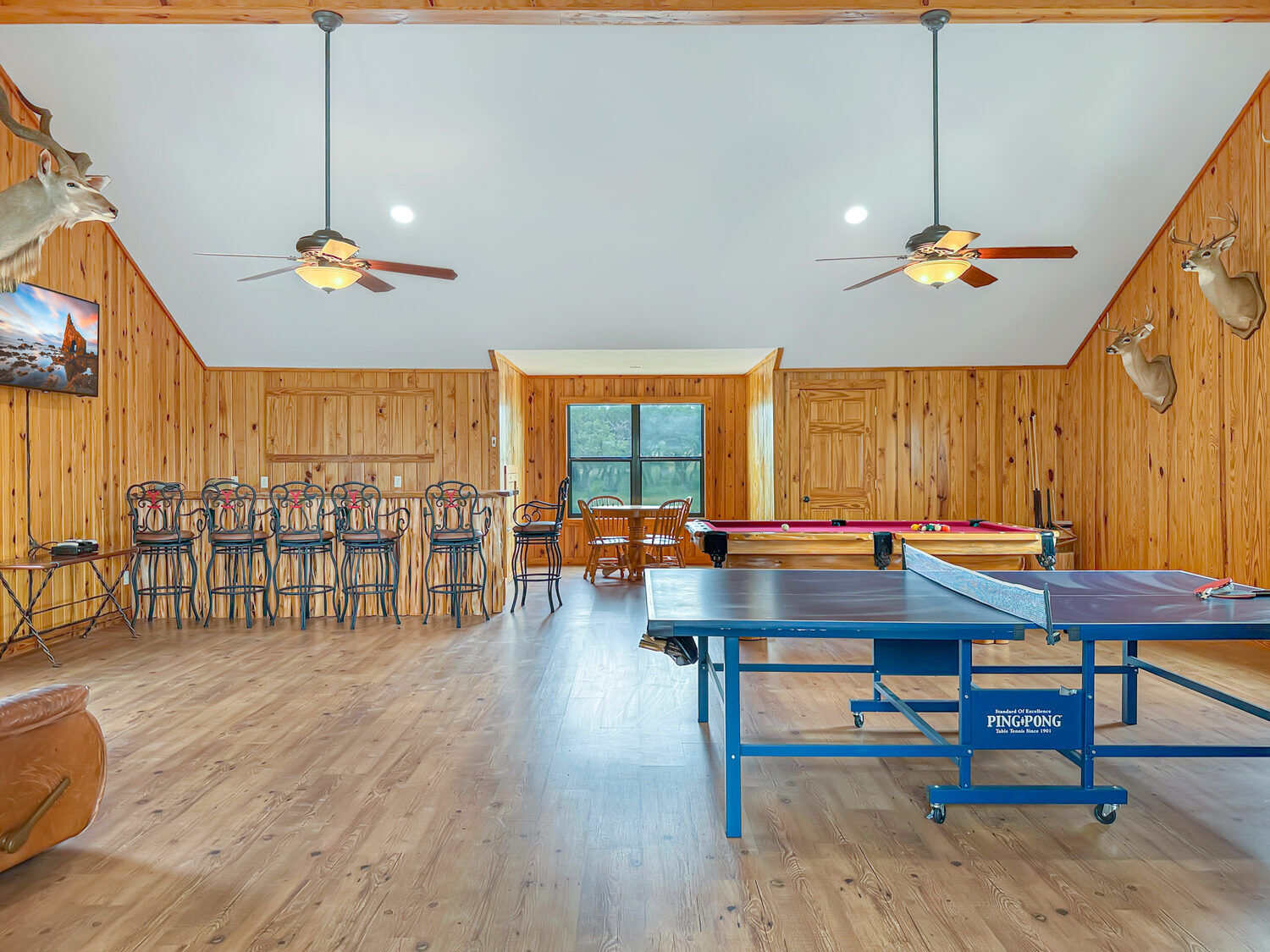 Upstairs game loft with a bar, stools, pool table and ping pong table under a vaulted knotty-pine ceiling