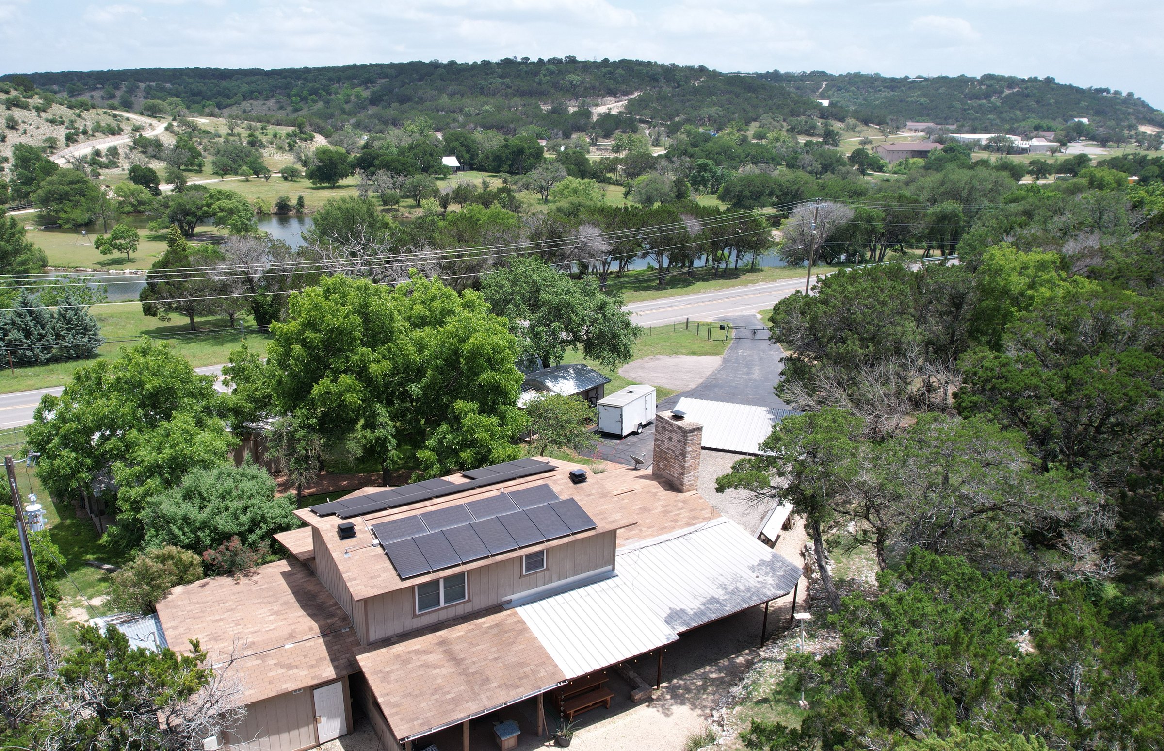 Aerial view of the house at 1406 N Harper Rd, Kerrville Texas, showing the new composition roof, the solar array, the outbuildings and the 2.57 wooded acres with the hill-country ridge behind