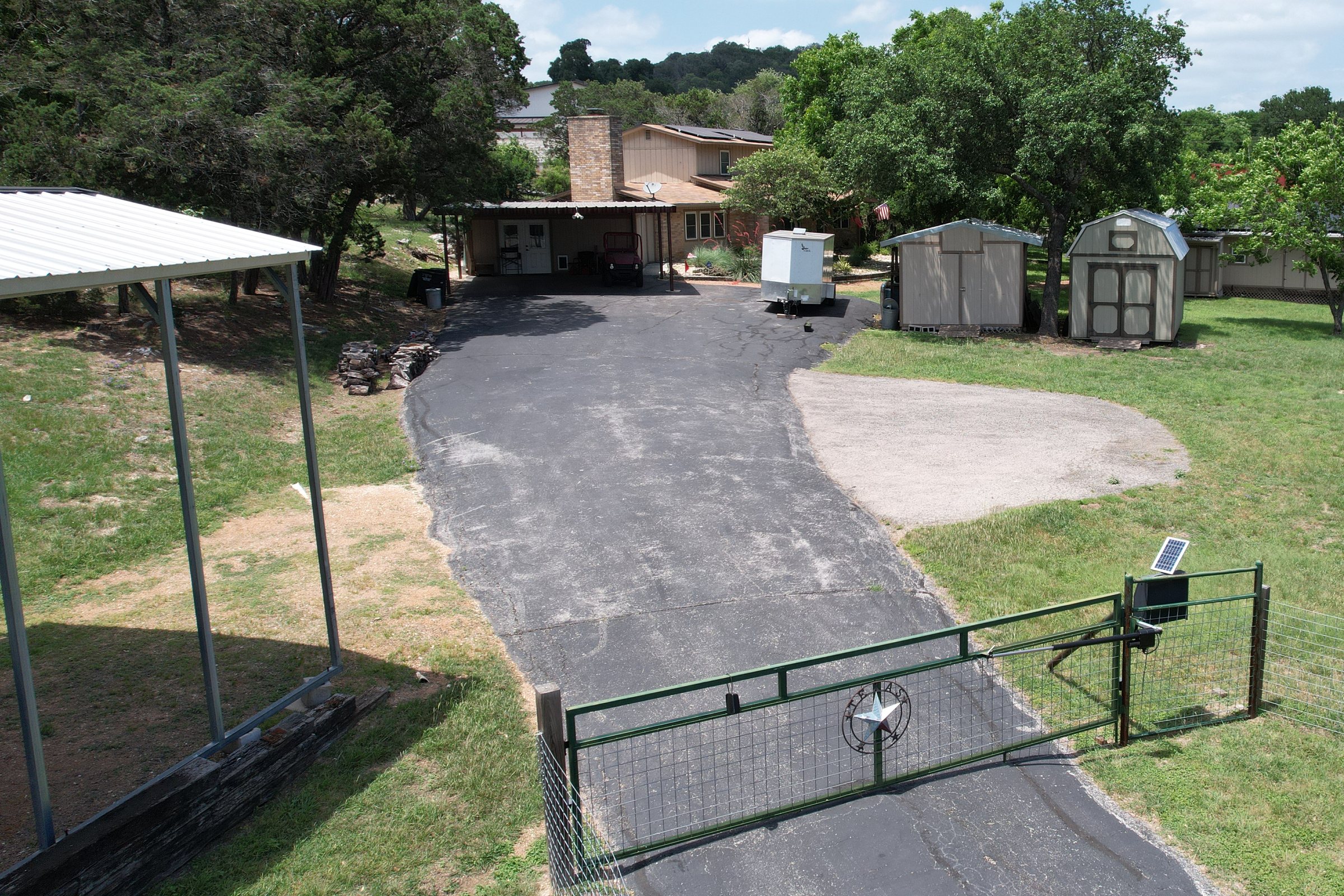 The homestead seen from the entrance gate — asphalt drive running back past the carport to the house, with both storage sheds on one side and the RV cover on the other