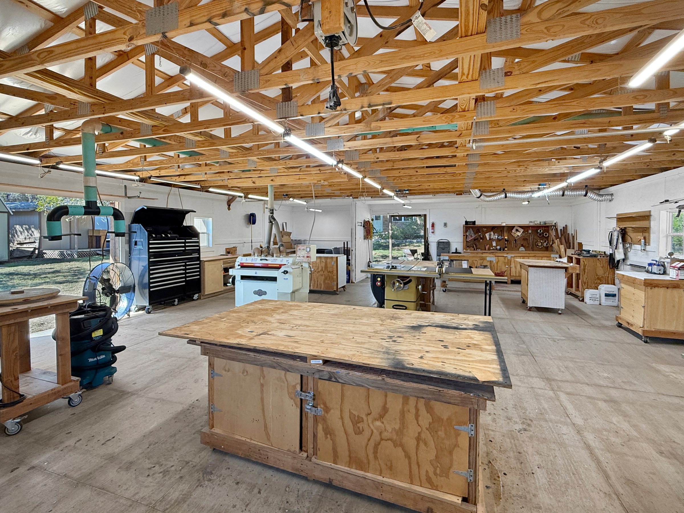 Inside the 30 by 38 foot detached workshop — open span under exposed wood trusses, run lighting, dust collection, workbenches and rolling tool stations on a concrete floor