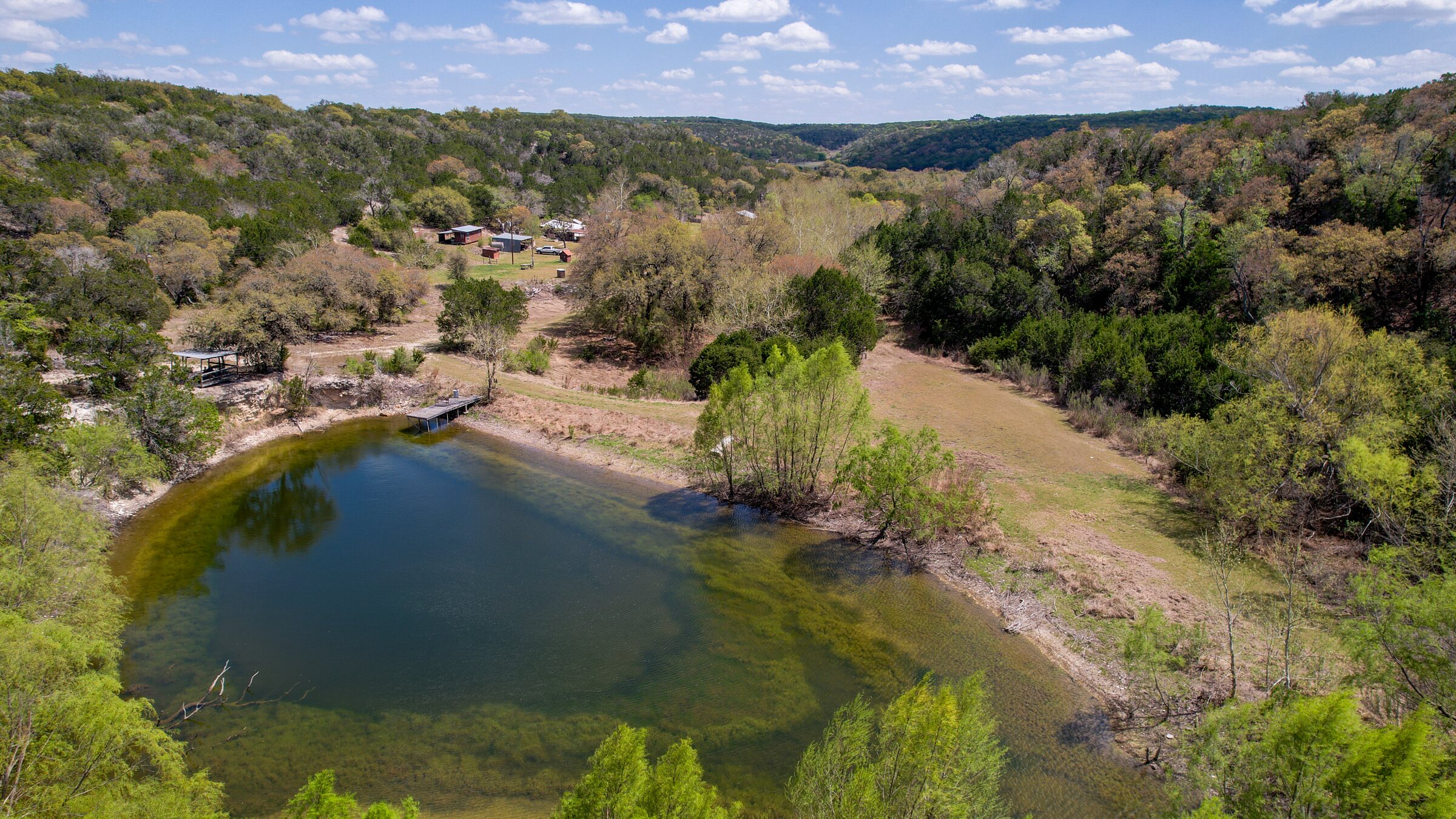 Aerial over the spring-fed pond at 150 Coker Hollow, Kerrville Texas, looking up the box canyon toward the homestead and the wooded hillsides beyond