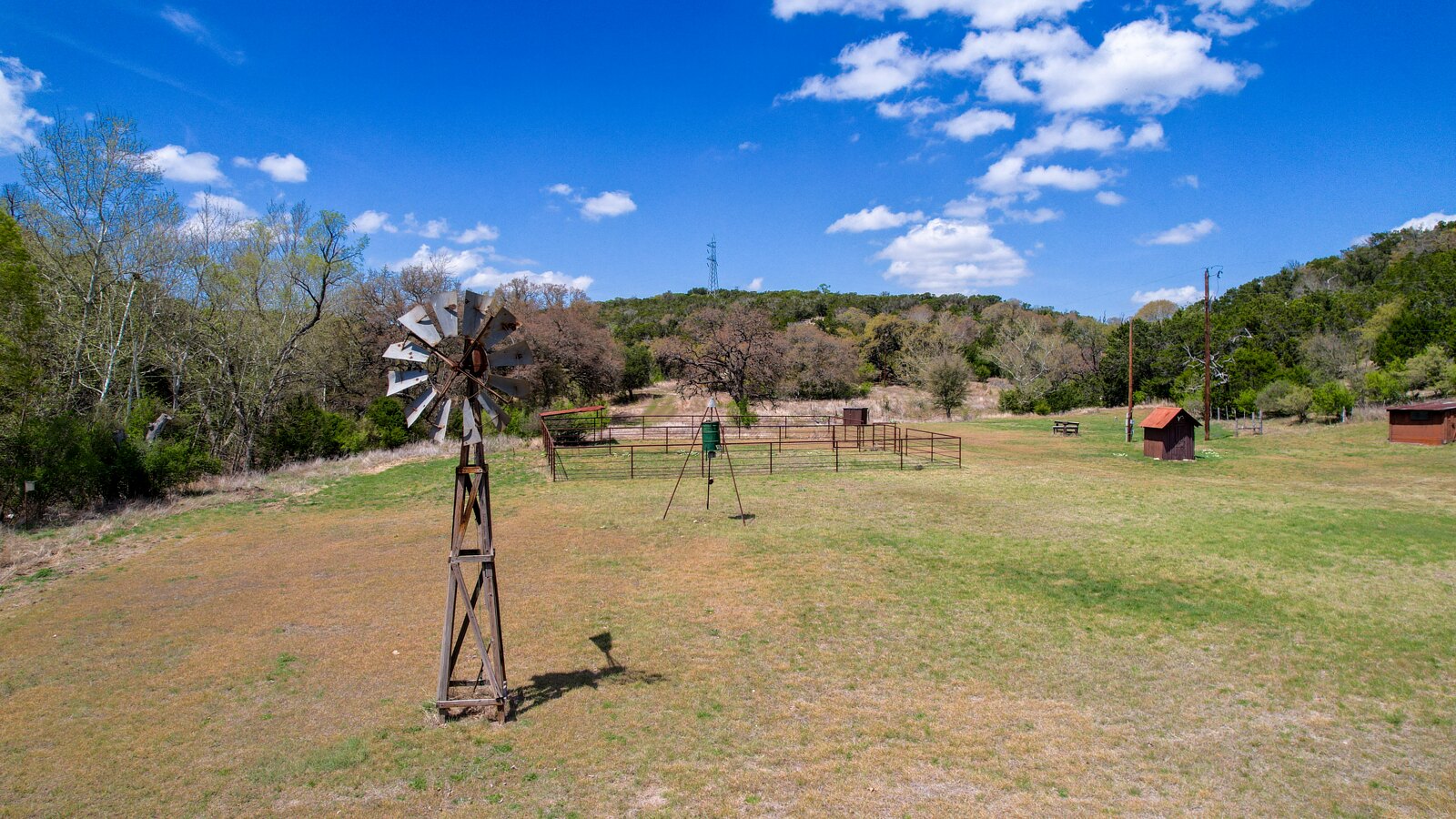 Open meadow at 150 Coker Hollow with a windmill in the foreground, corrals and a game feeder behind it, and wooded hills rising at the back