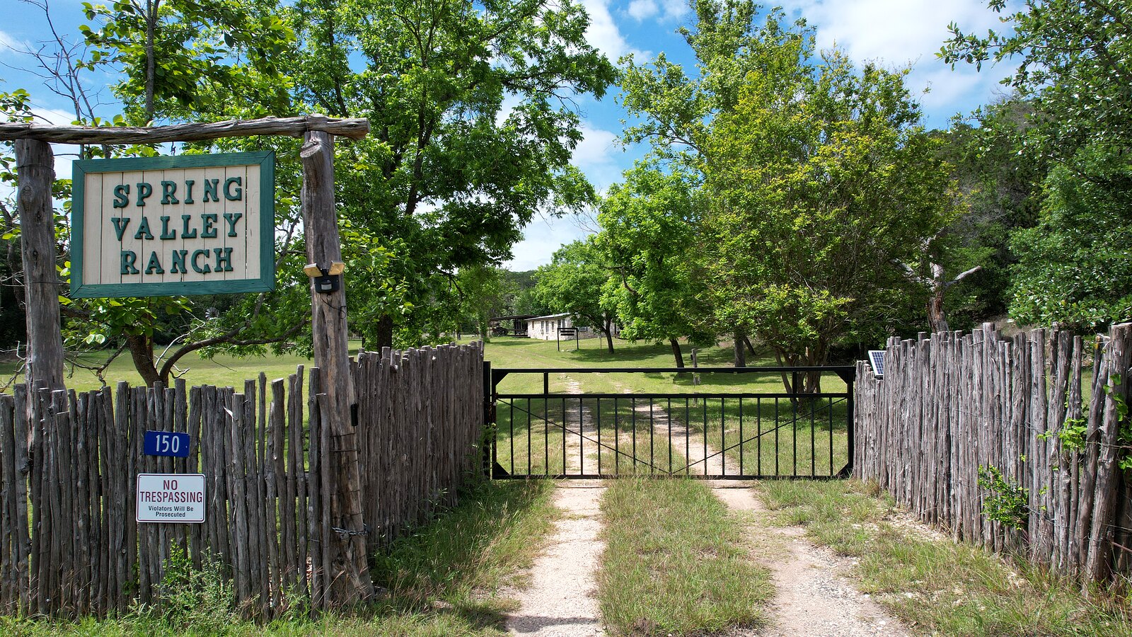 The entrance gate at 150 Coker Hollow with the Spring Valley Ranch sign on a cedar-post header, the 150 address plate, cedar picket fencing and the caliche drive running in past the gate