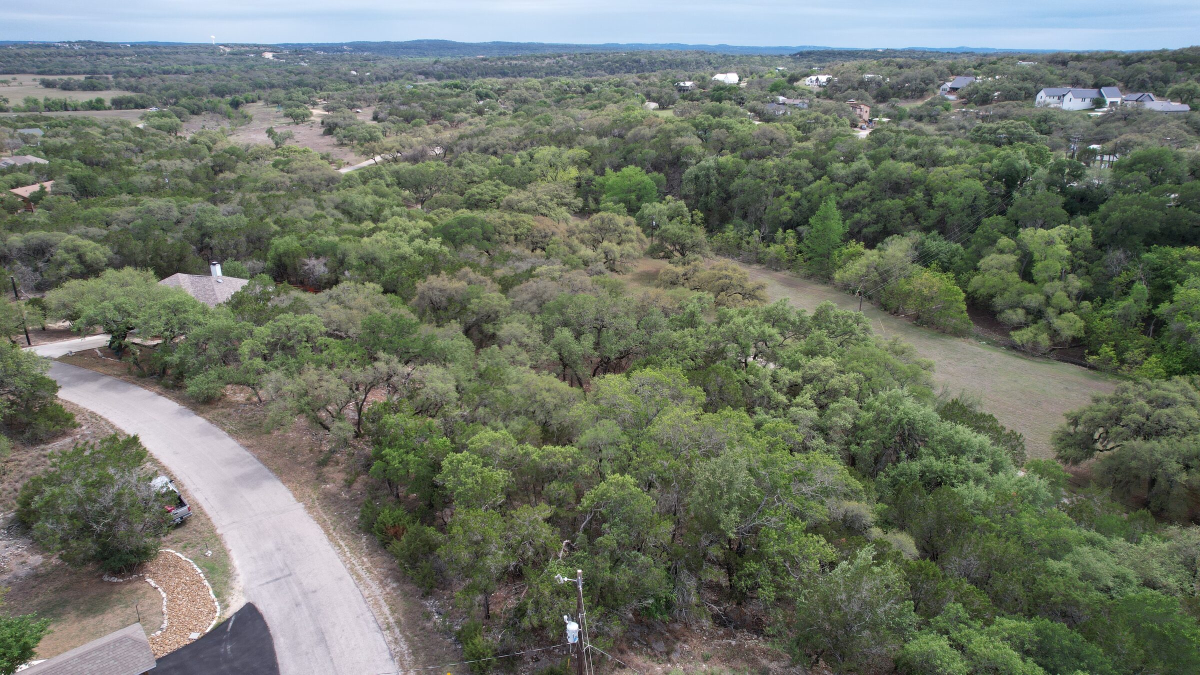 Drone view over the wooded lot at 183 Palm, Spring Branch, Texas — oak canopy in the foreground, open ground falling away to the right, and a Hill Country ridgeline across the top of the frame