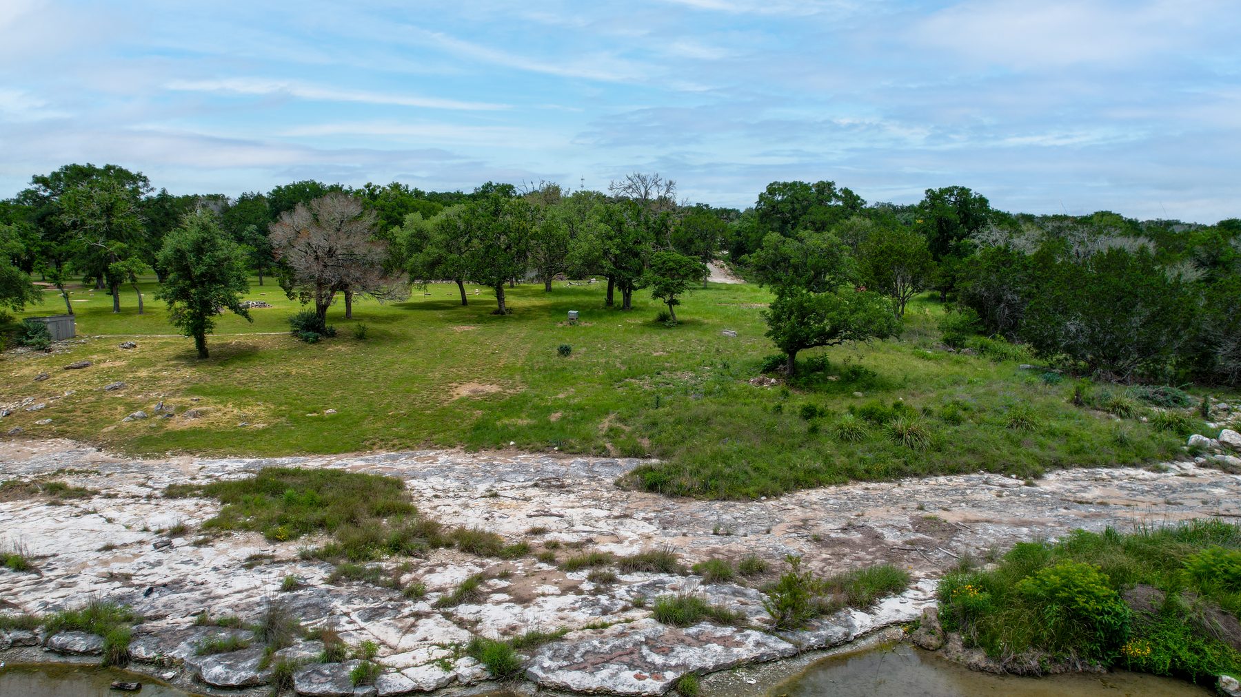 Aerial view over live oaks and open grass toward the river, with a flat limestone shelf in the foreground