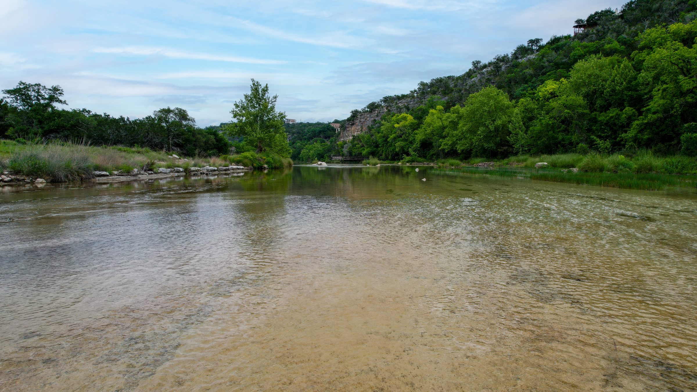 Aerial view over shallow, clear water on the North Fork of the Guadalupe River, with a wooded limestone bluff rising on the right under a blue sky