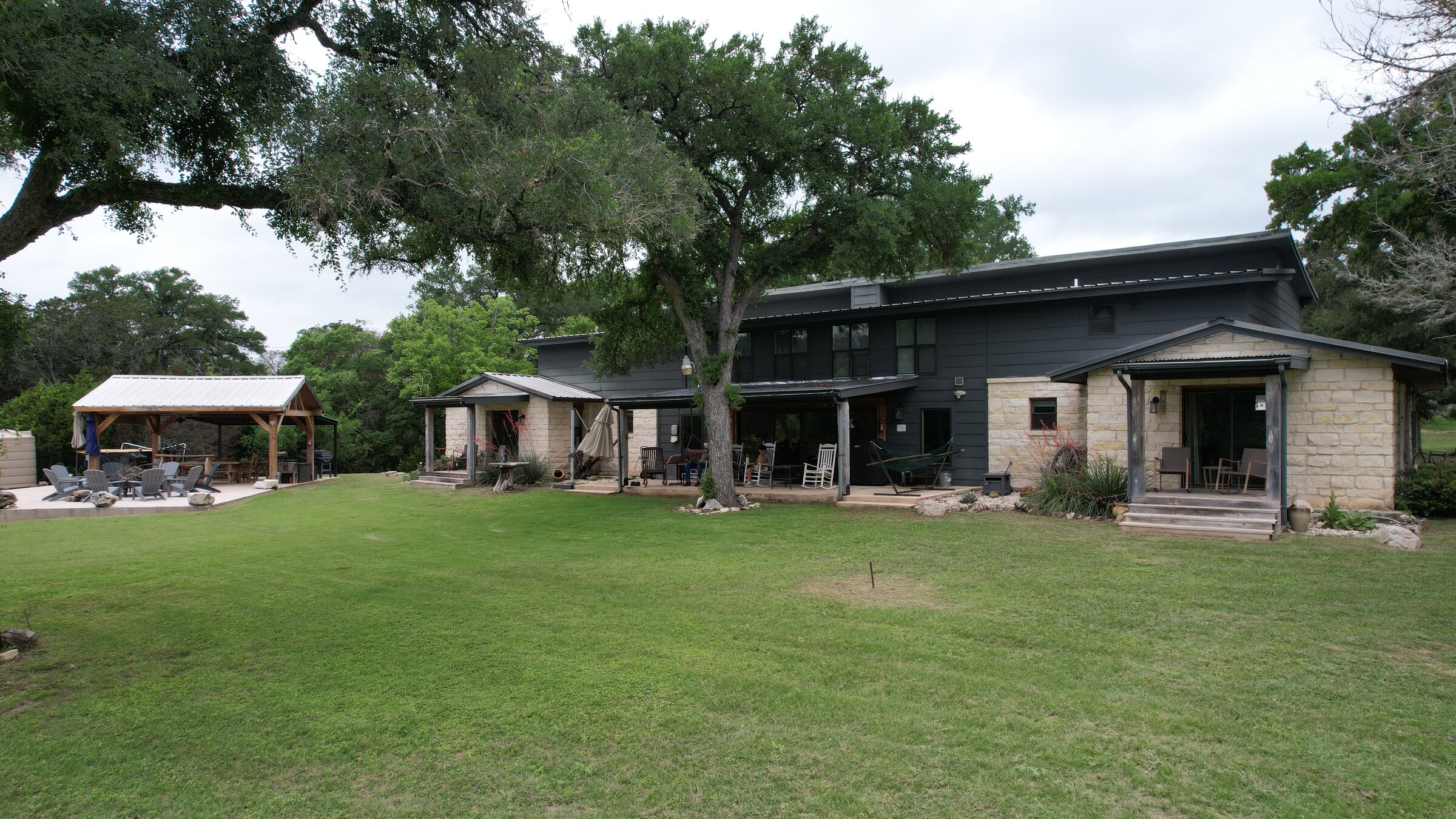 The house at 196 Rock Bottom Road, Hunt Texas, seen across the lawn — two stories in dark siding and native rock under a metal roof, with covered porches and the timber-frame outdoor kitchen off to the left under mature live oaks