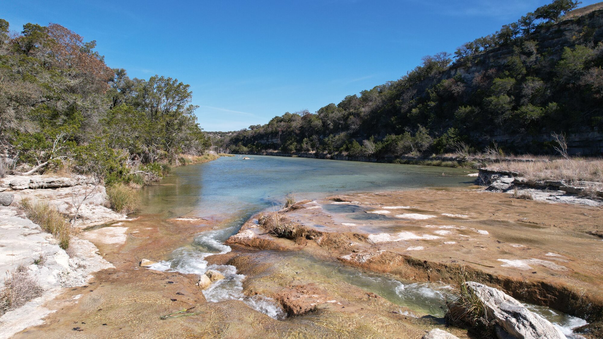 The North Fork of the Guadalupe at 196 Rock Bottom Road — clear green water running over a wide limestone shelf, with wooded banks and the bluff line rising on the far side