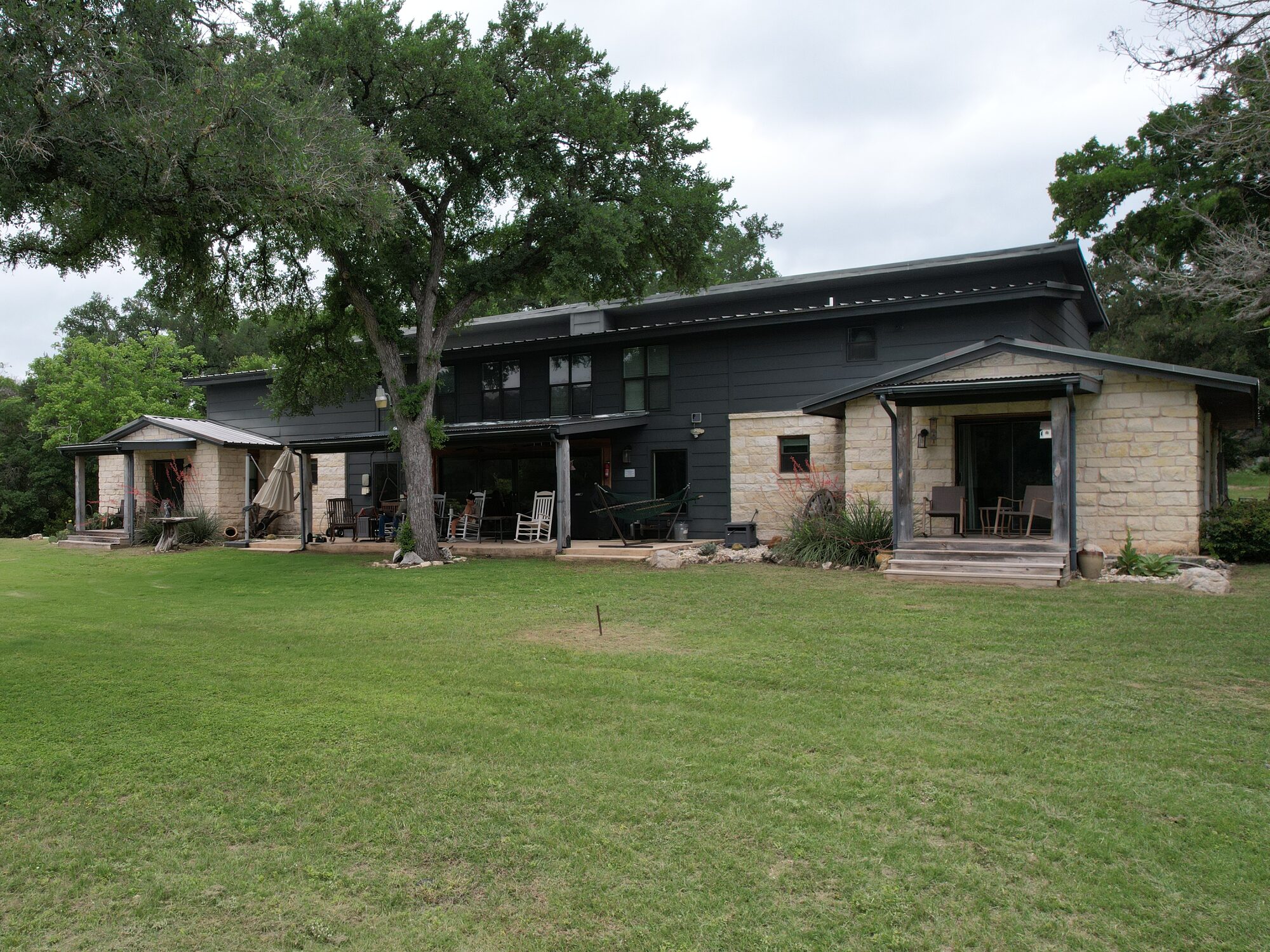 The house at 196 Rock Bottom Road from the lawn — two stories in dark siding and native rock under a metal roof, with covered porches running the length of it and a live oak in front