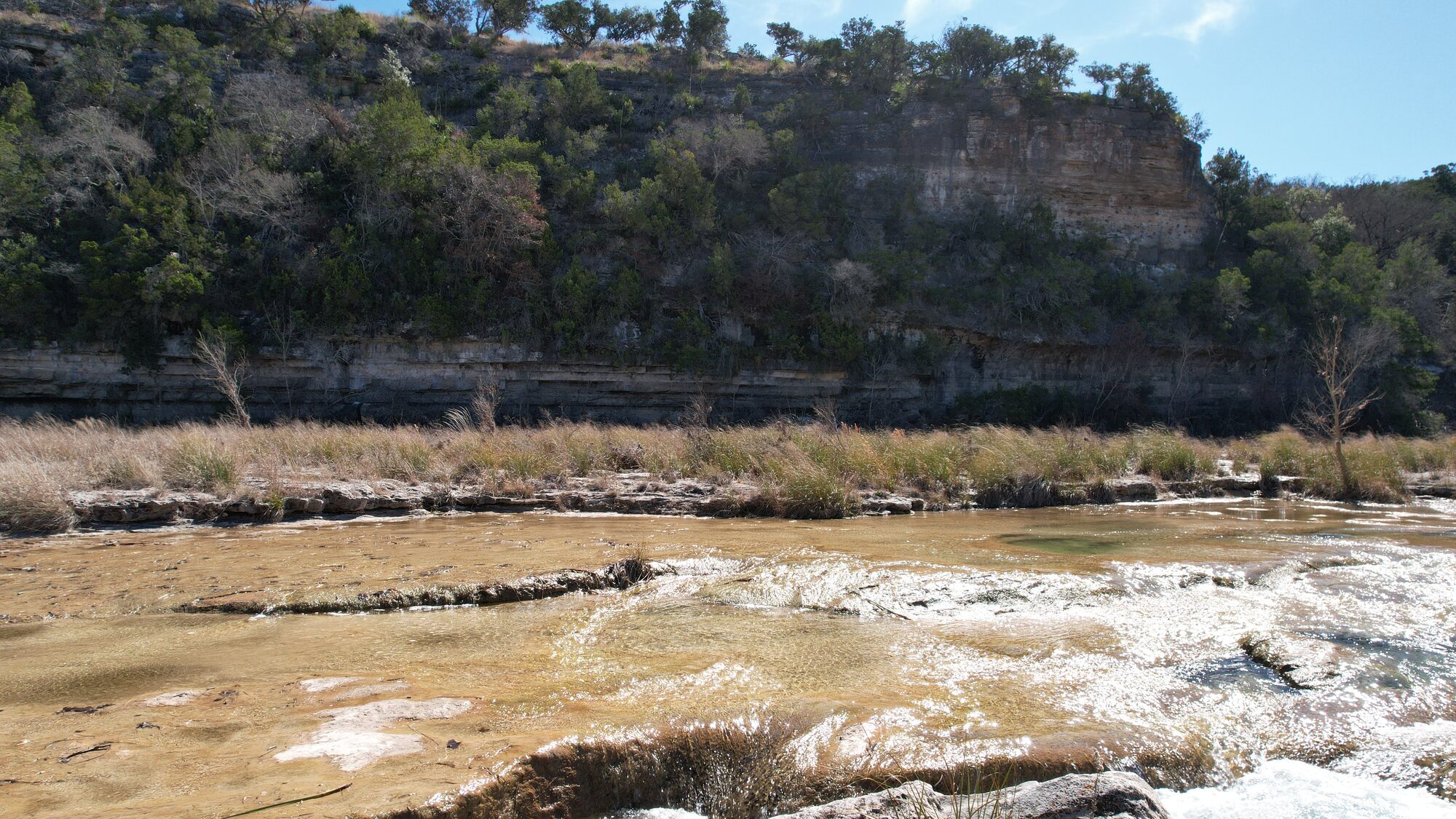 The layered limestone bluff above the North Fork of the Guadalupe at 196 Rock Bottom Road, with cedar and oak growing along the ledges and the rock riverbed in the foreground