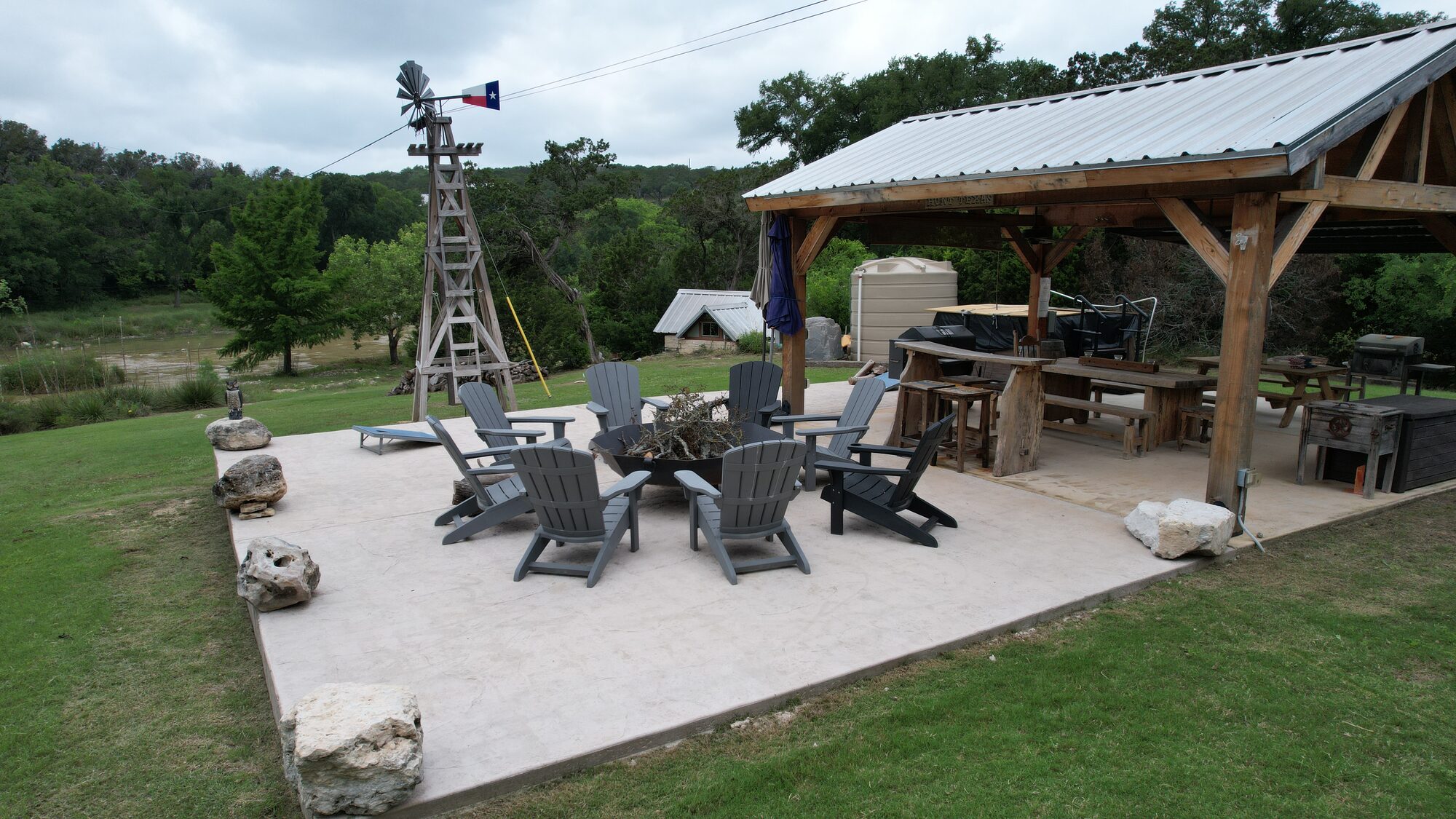 The fire pit patio at 196 Rock Bottom Road — a poured patio edged in limestone boulders with Adirondack chairs around a fire ring, a wooden windmill flying a Texas flag, and the timber-frame outdoor kitchen under a metal-roofed pavilion to the right