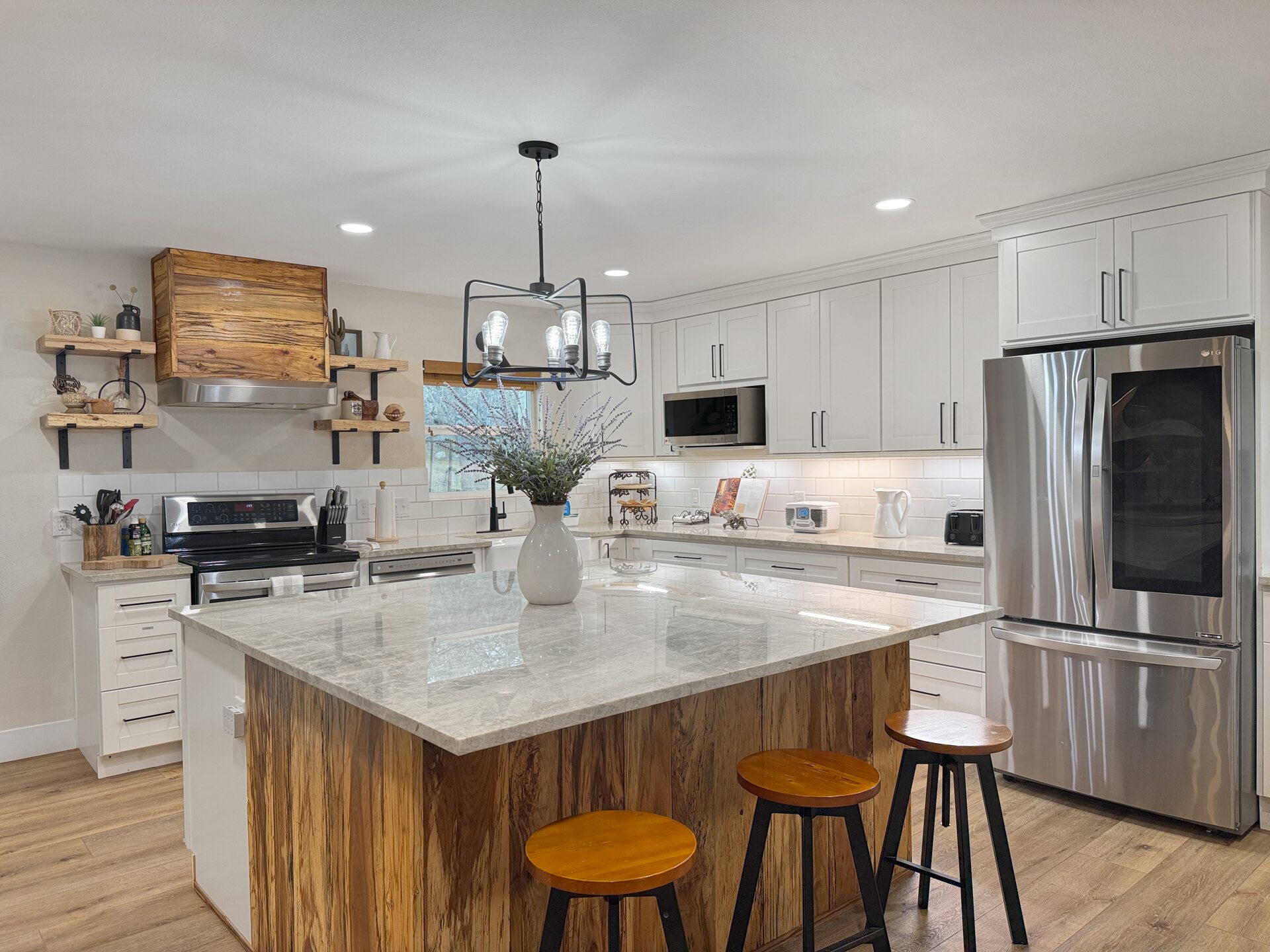 The kitchen at 196 Rock Bottom Road — a large quartzite-topped island with a live-edge wood base and bar stools, white shaker cabinetry, subway tile, stainless appliances and a timber range hood