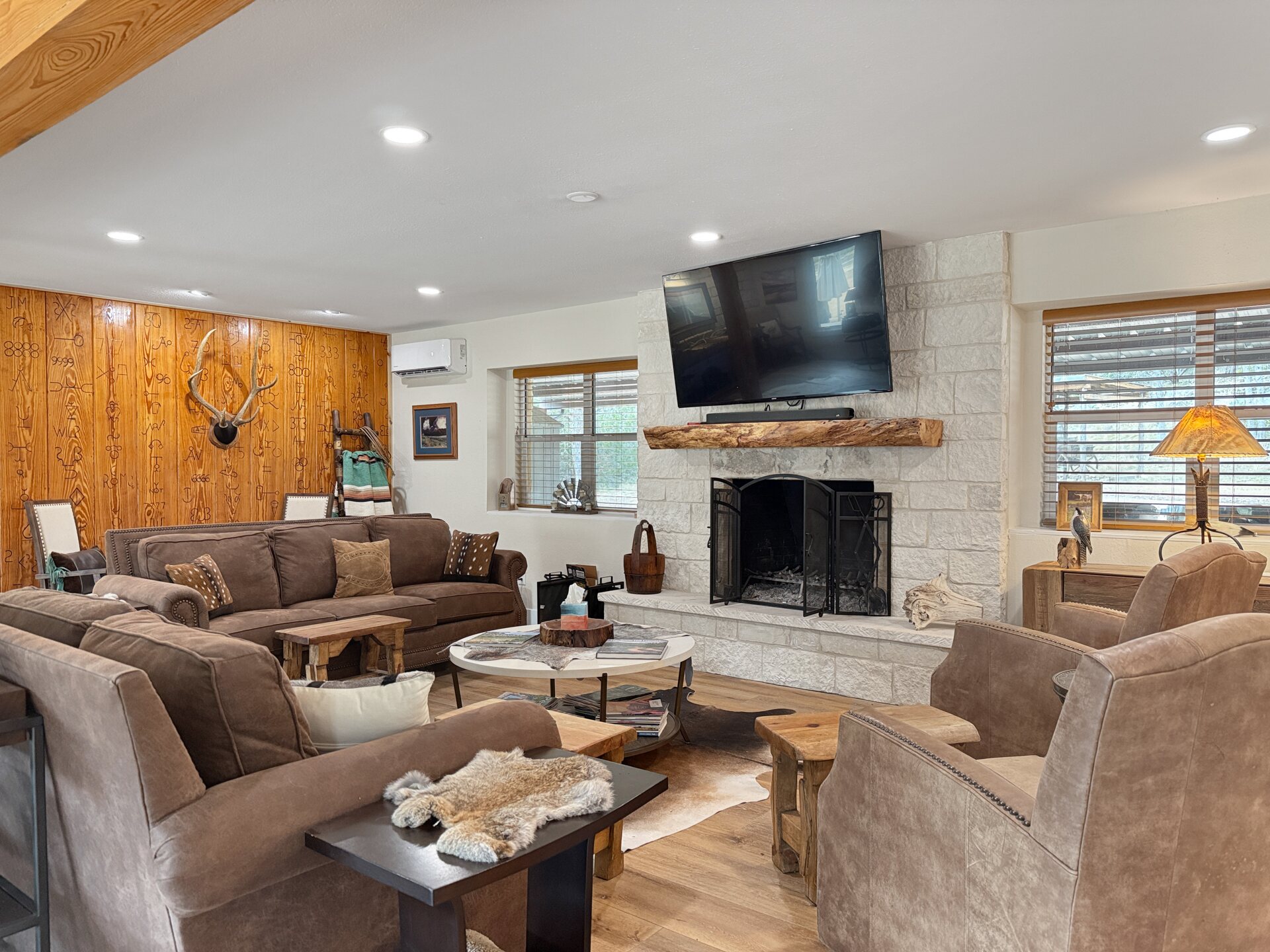 The great room at 196 Rock Bottom Road — a limestone fireplace with a live-edge timber mantel, exposed beam overhead, knotty pine wall, leather seating and a mini-split head on the wall