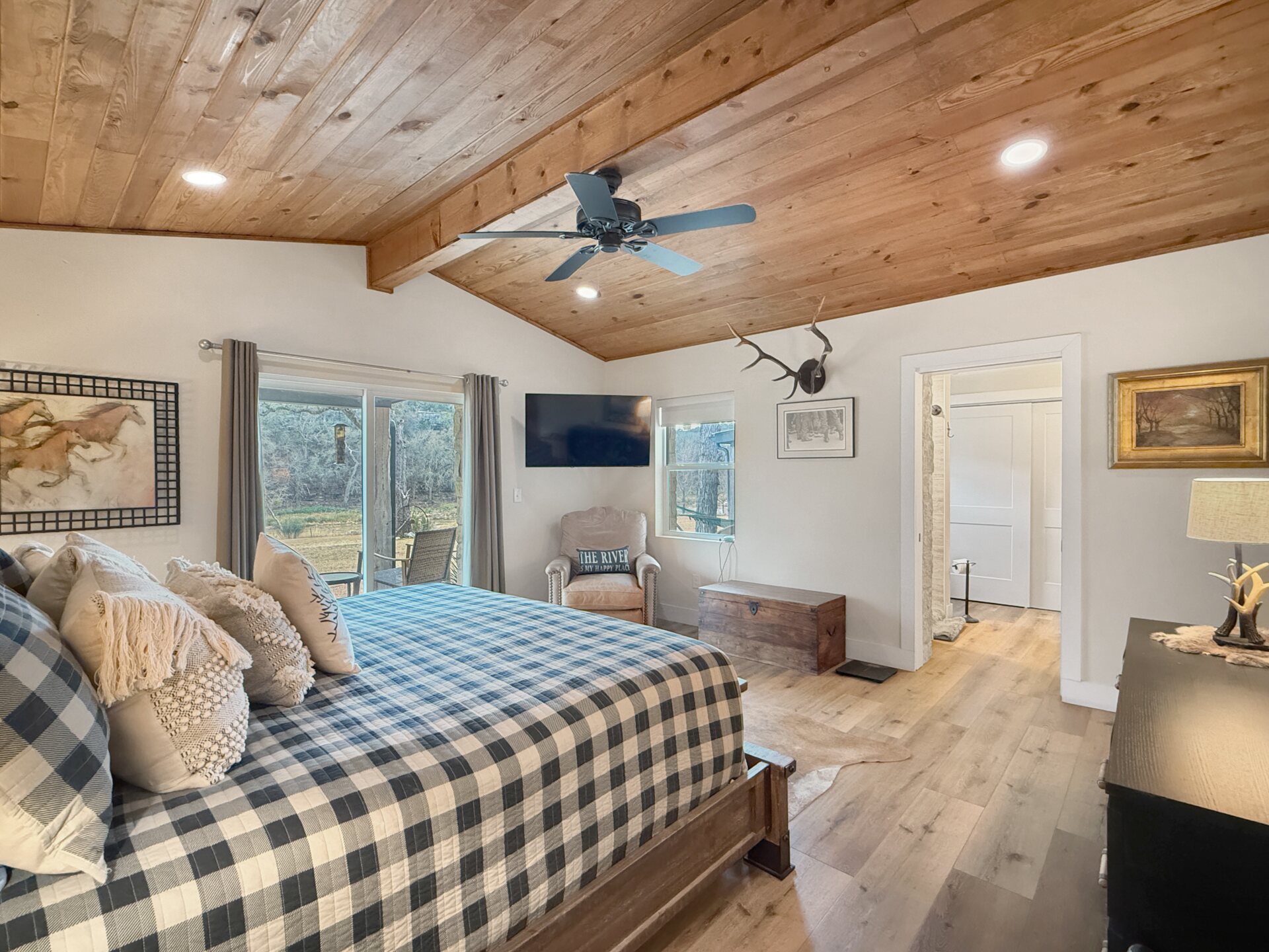 The master suite at 196 Rock Bottom Road — vaulted tongue-and-groove wood ceiling with an exposed beam and ceiling fan, wide-plank floors, a king bed and a sliding door out to the deck