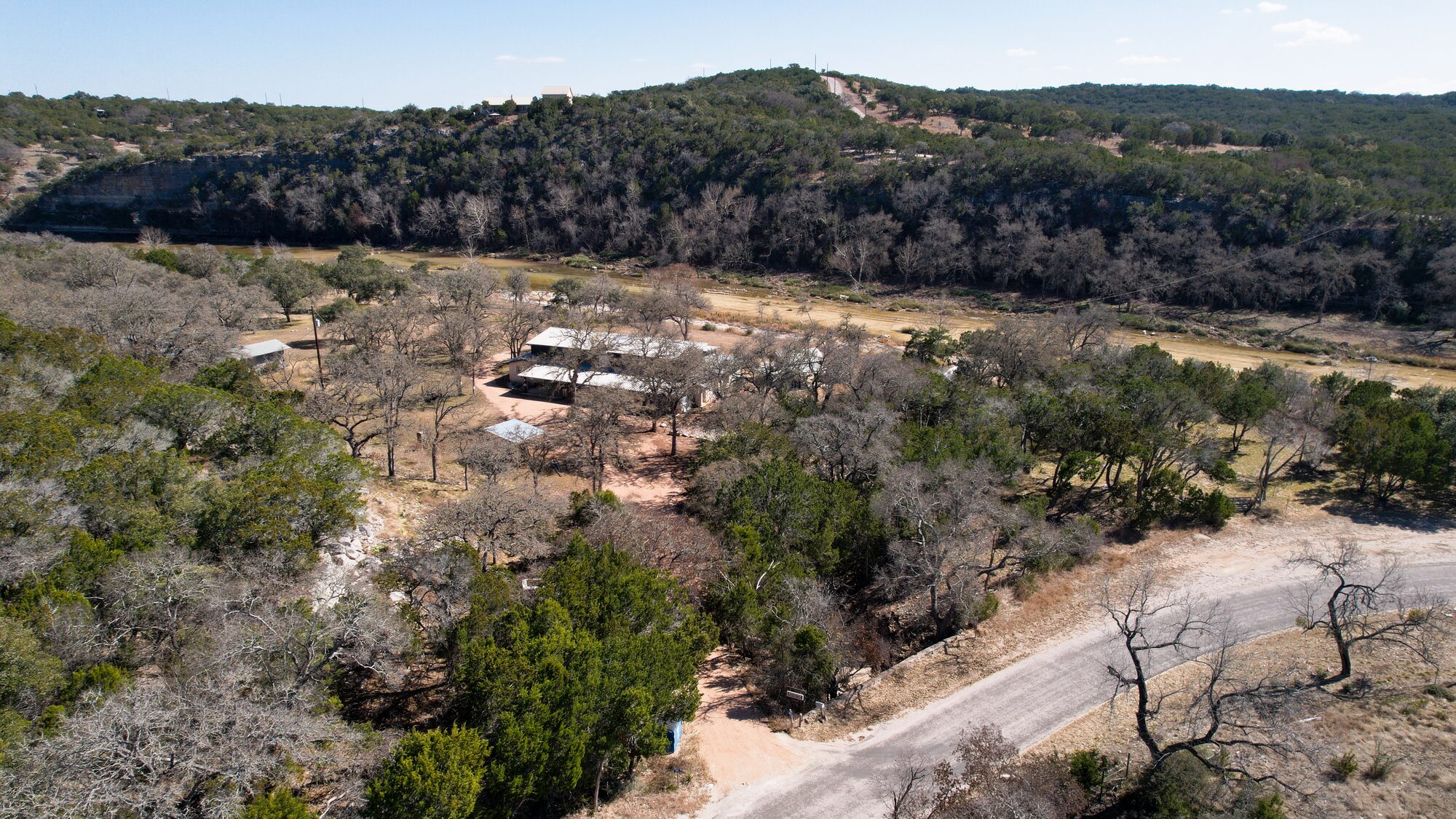 Aerial over 196 Rock Bottom Road showing the improvements among the oaks in the middle of the frame, the river bottom running across behind them, the county road curving through the foreground and the bluff line beyond