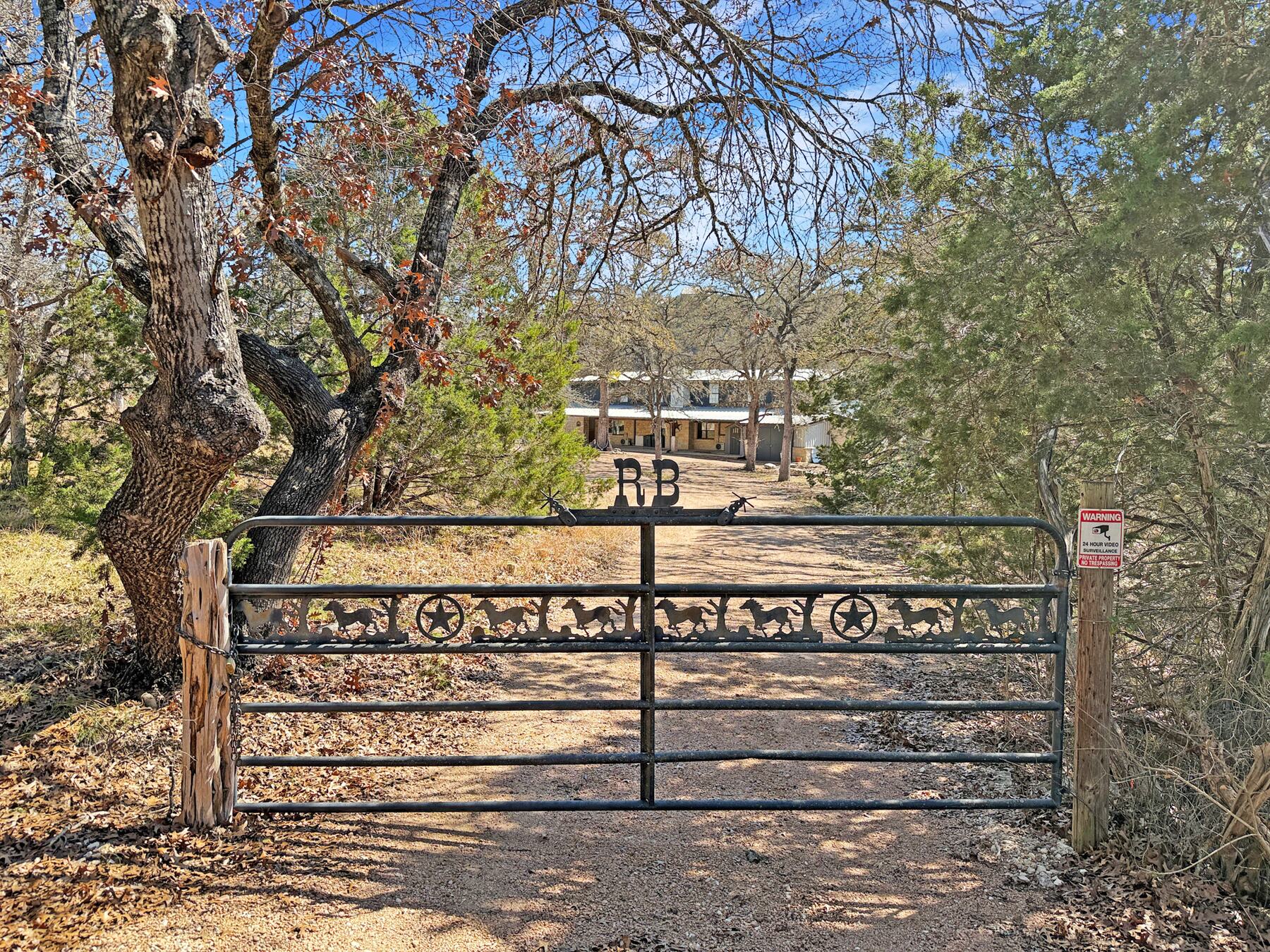 The custom iron ranch gate at 196 Rock Bottom Road, with running-horse and star ironwork across the panels, cedar posts either side and the caliche drive running in toward the house