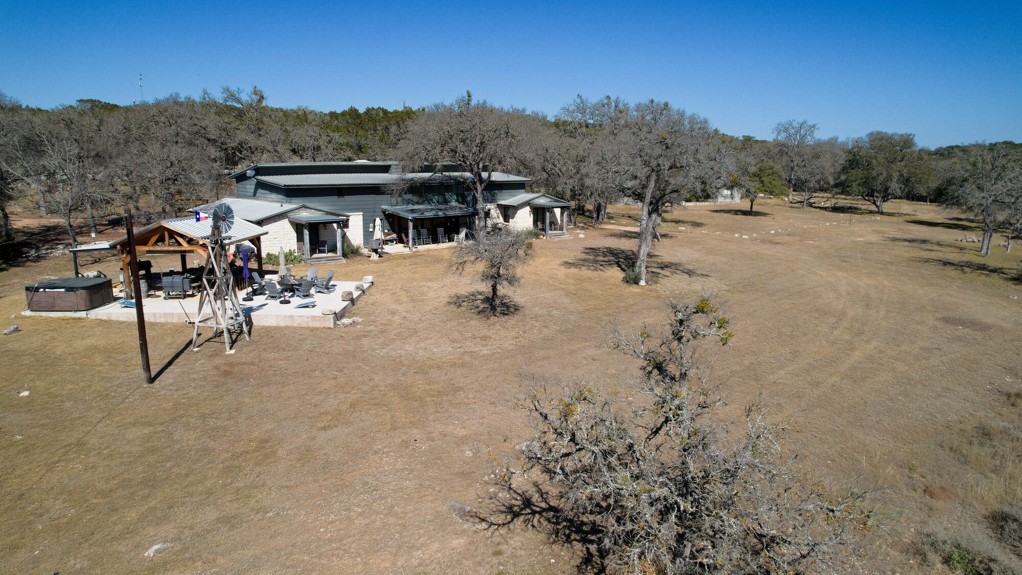 Aerial across 196 Rock Bottom Road showing the house, the metal-roofed pavilion, the windmill and the fire pit patio on the near side, with open ground and live oaks running out behind