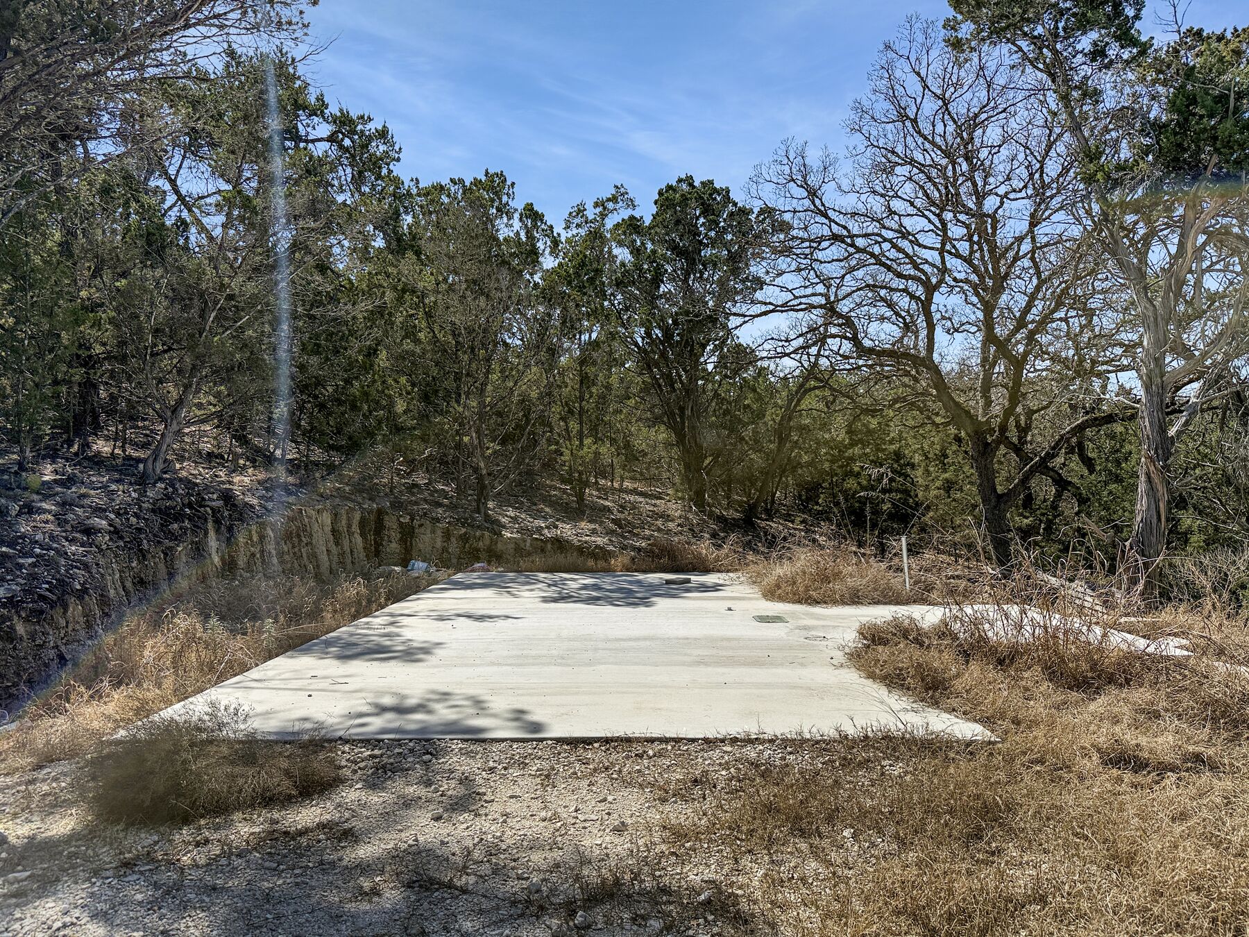 The prepared concrete pad at 214 Hazelett Dale Rd photographed from ground level, poured against a cut limestone bank with dormant grass and bare trees behind it