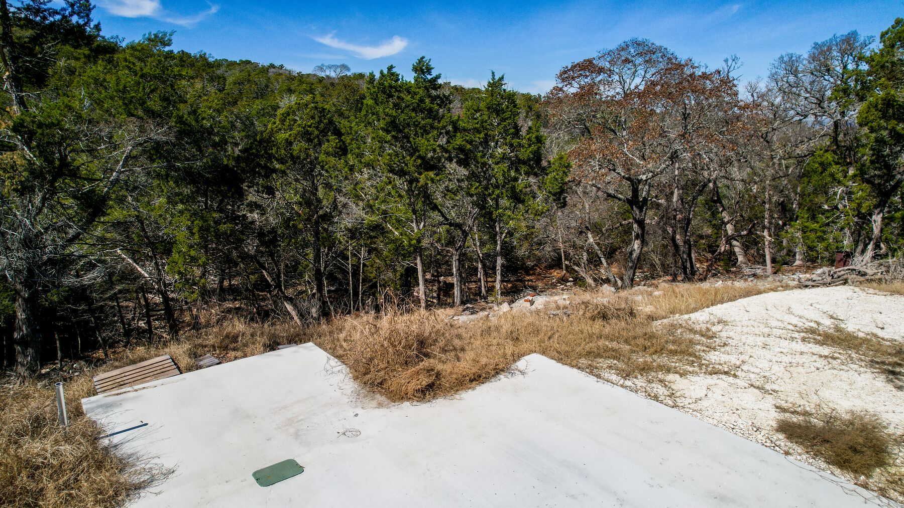Aerial over the prepared concrete pad at 214 Hazelett Dale Rd, the slab filling the lower half of the frame with dormant gold grass around it and wooded hills rising beyond