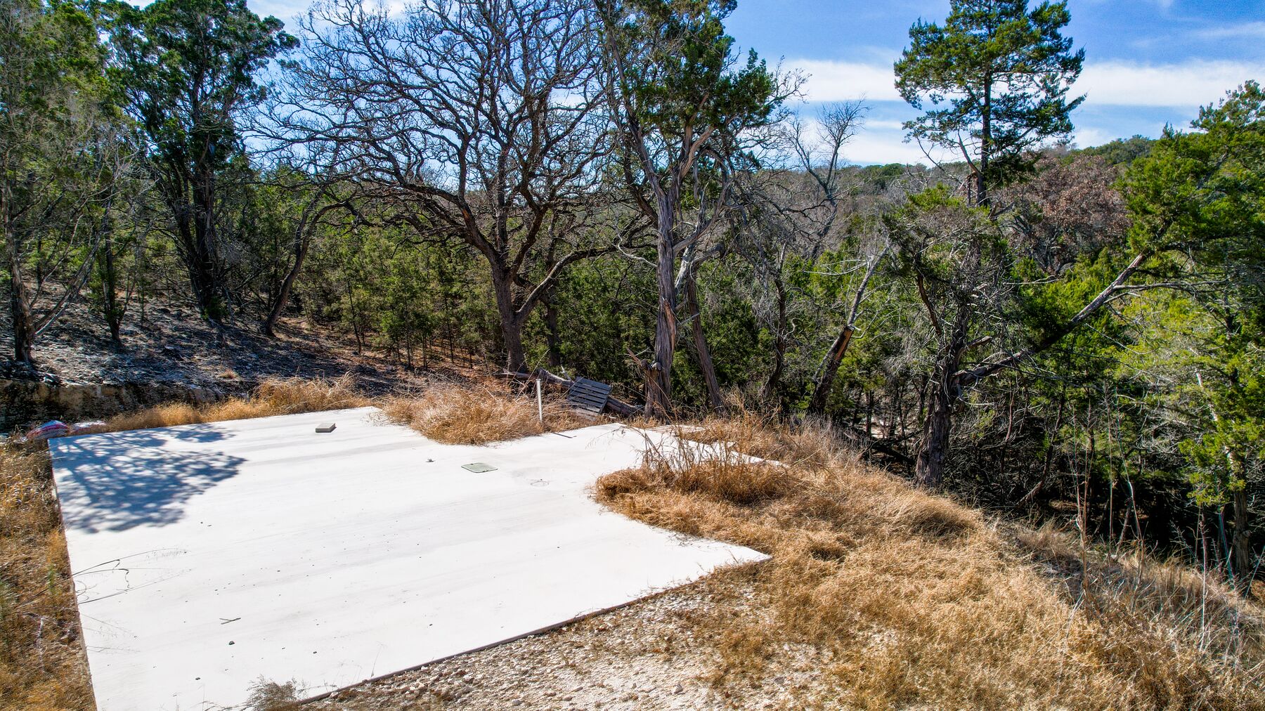 Wide aerial over the building site at 214 Hazelett Dale Rd, the cleared ground in the middle of the frame and a Hill Country ridge line visible past the treeline at the right
