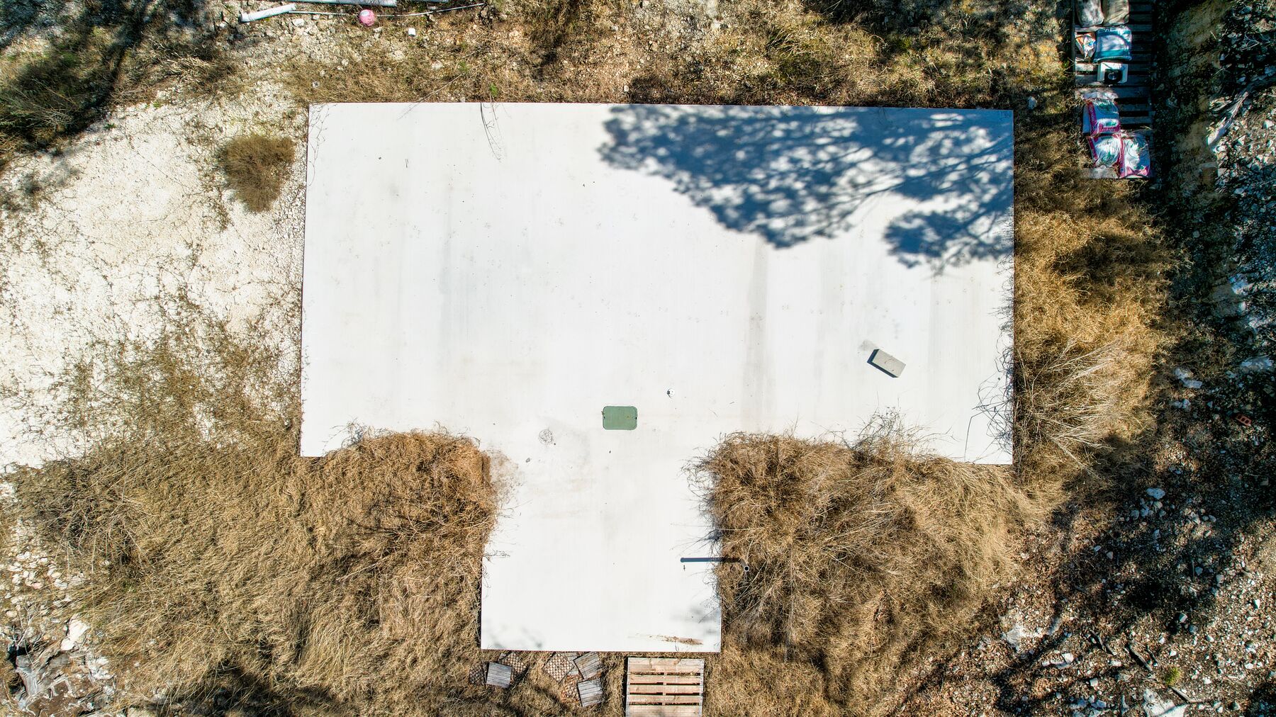 Nadir drone frame straight down over the prepared pad at 214 Hazelett Dale Rd, the poured slab and the pale caliche apron around it set into the surrounding tree cover
