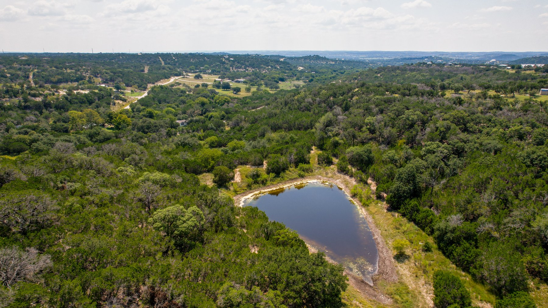 Oblique aerial of the pond at 230 Marshall Dr set among cedar and live oak, with the valley and open pasture running out beyond