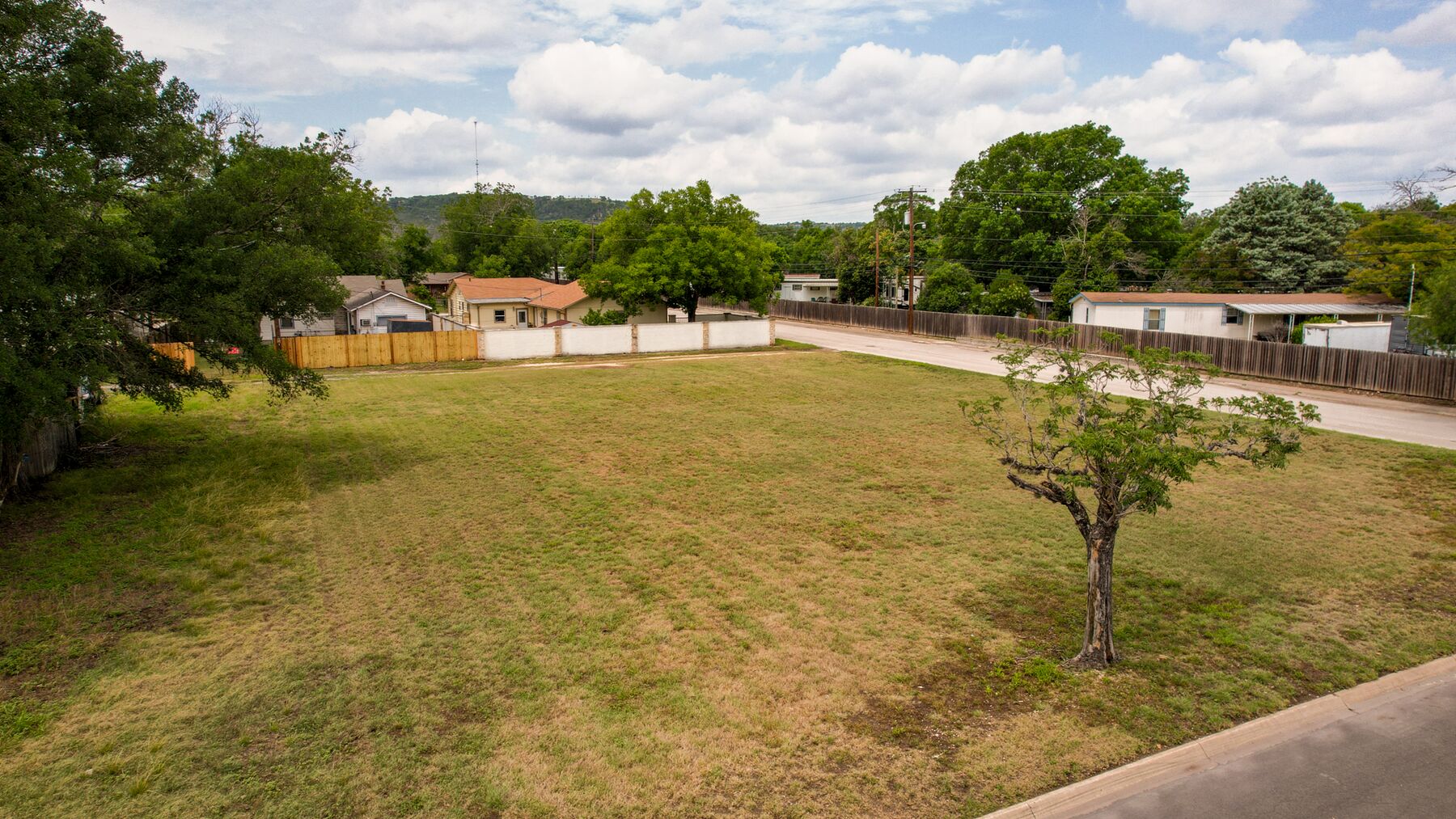 Elevated view across the property from the street side. The ground is level and open, with mown grass throughout and a single mature tree standing near the pavement.