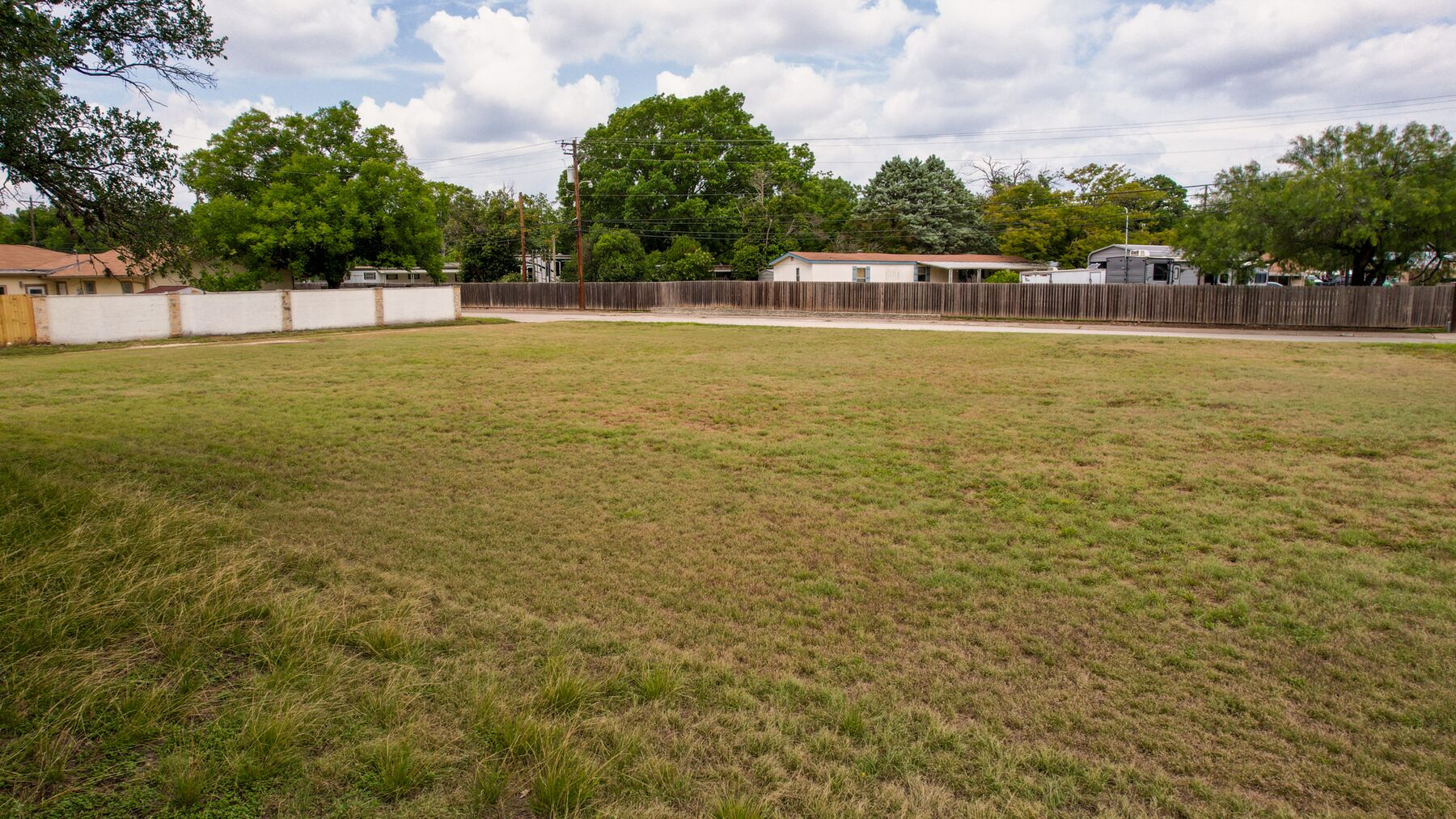 Ground-level view across the open grass toward the paved street on the far side. A white masonry wall runs along the left-hand edge; the fences and buildings beyond the street are on adjoining property.