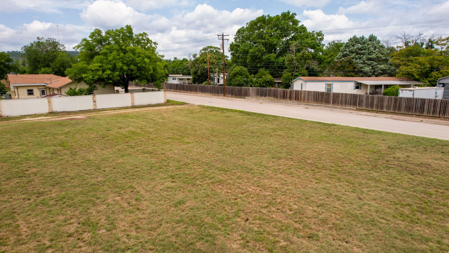 Wider view with the masonry wall along the left and the street running along the right. The ground is clear and grassed, with no structures standing on it.