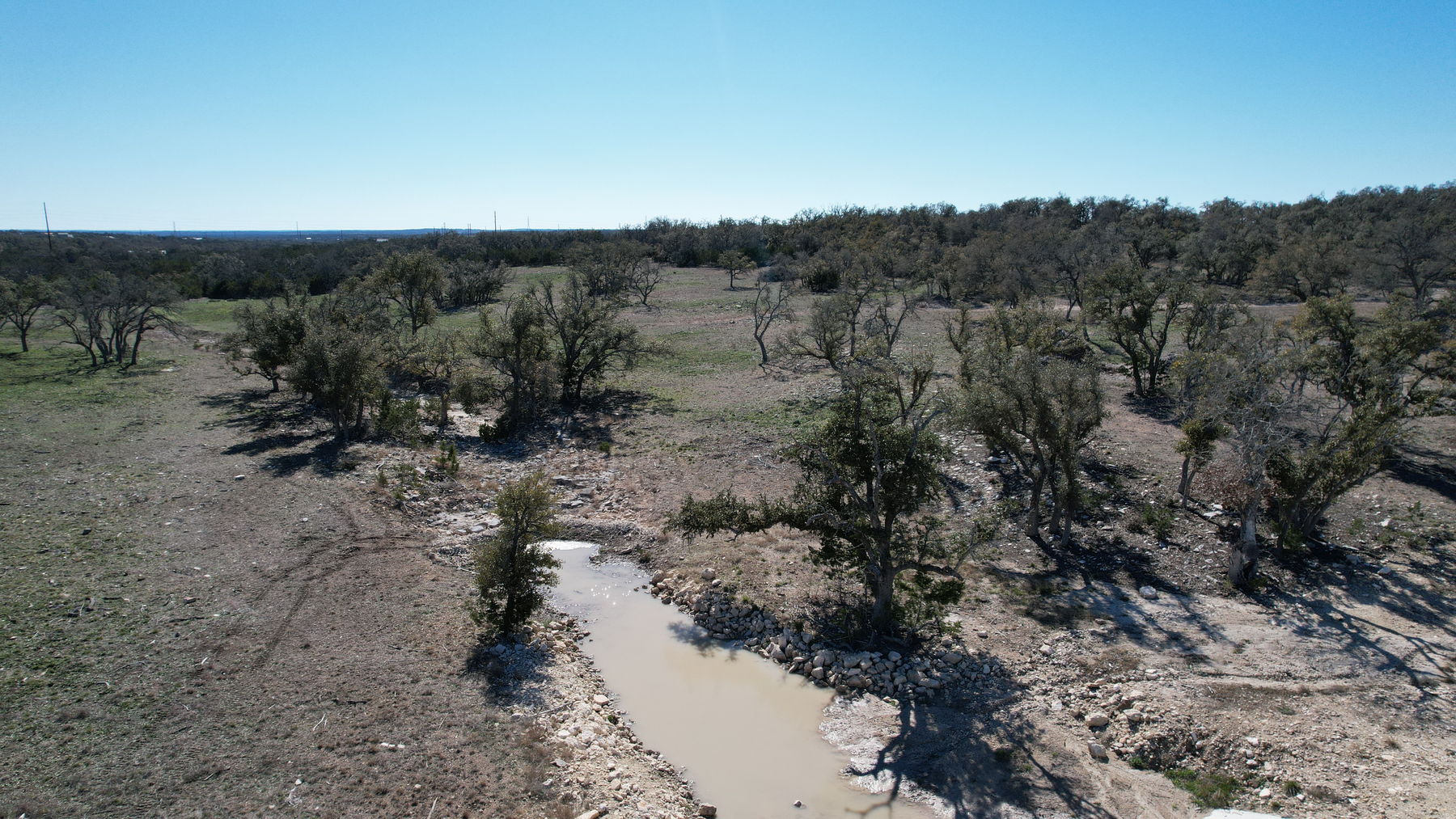 Aerial of the drainage at the low point, its channel edged with loose stone, holding shallow silty water