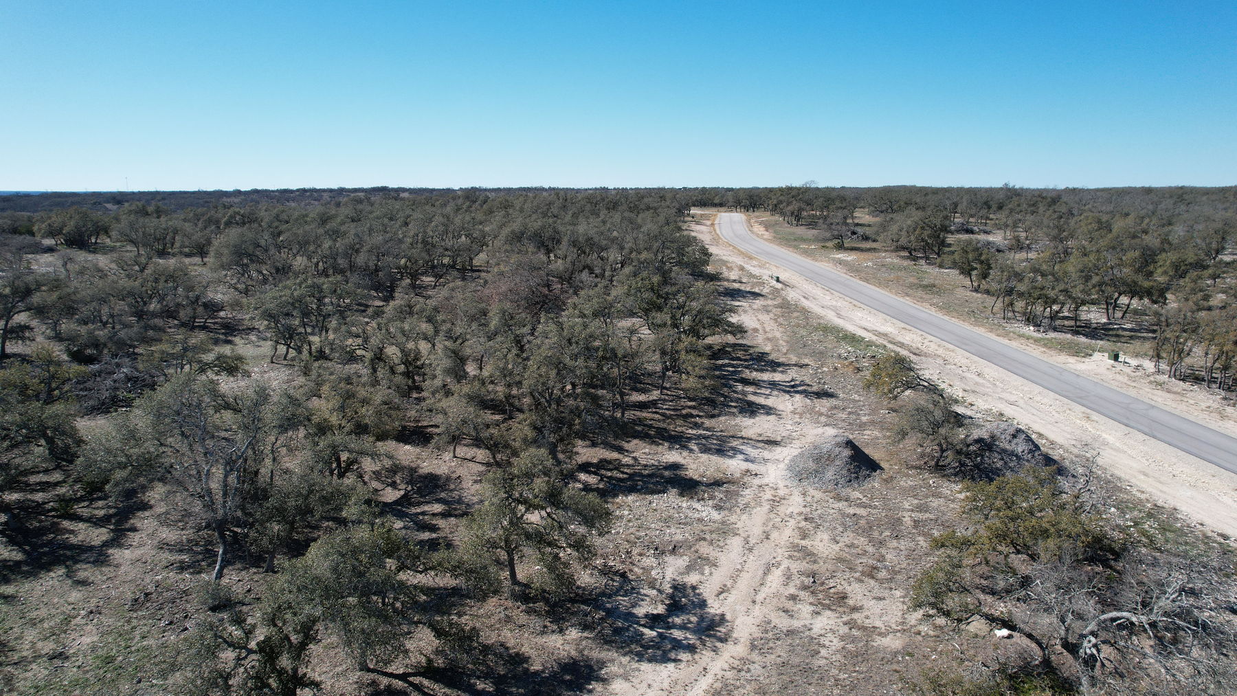 Aerial of the paved subdivision road running alongside the tree cover, with a pale mound of stockpiled road material at right