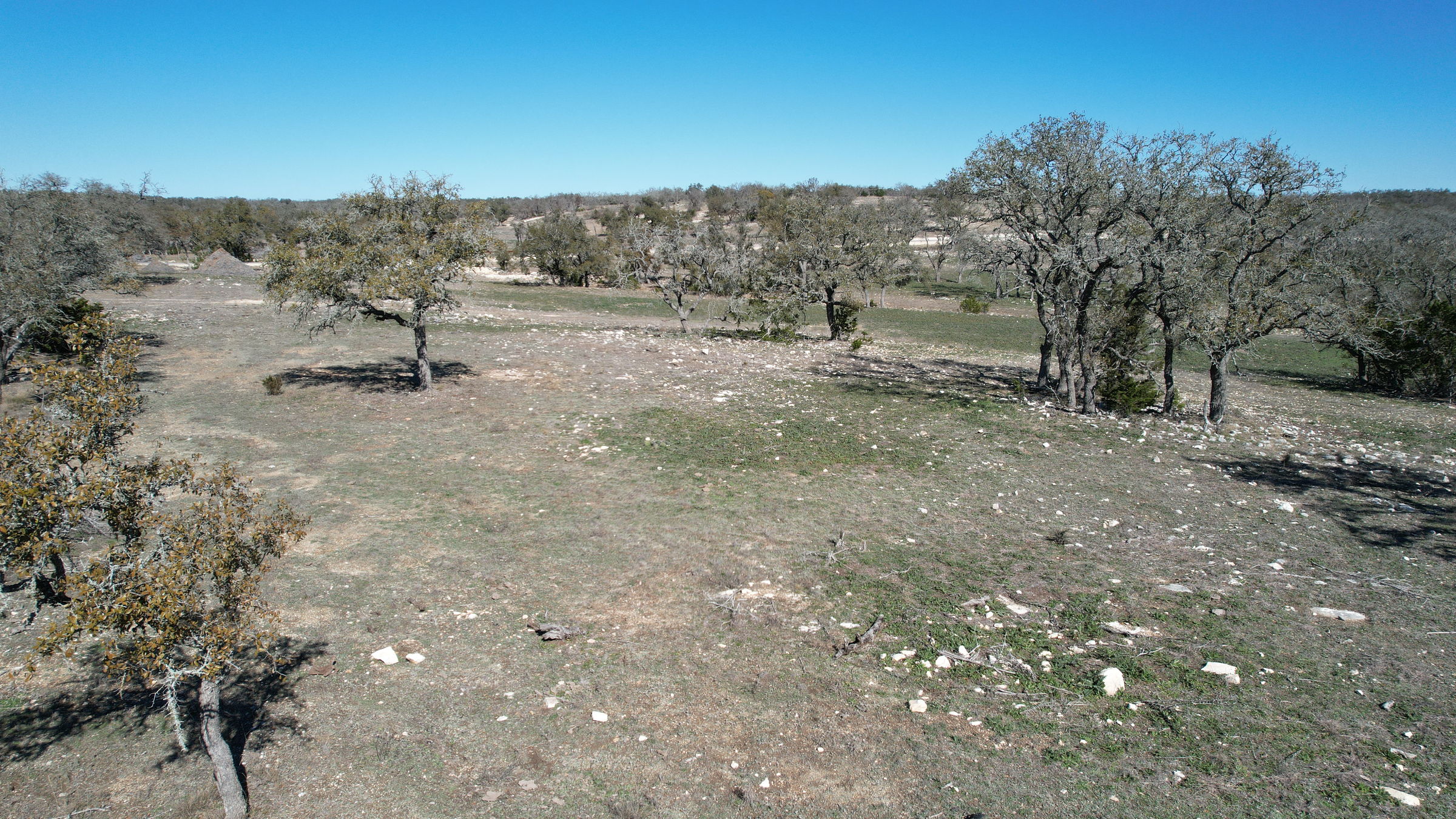 Low aerial across open ground at Lot 34 N Brooklyn Dr, with scattered mature live oaks, limestone breaking the surface and Hill Country beyond under a clear winter sky